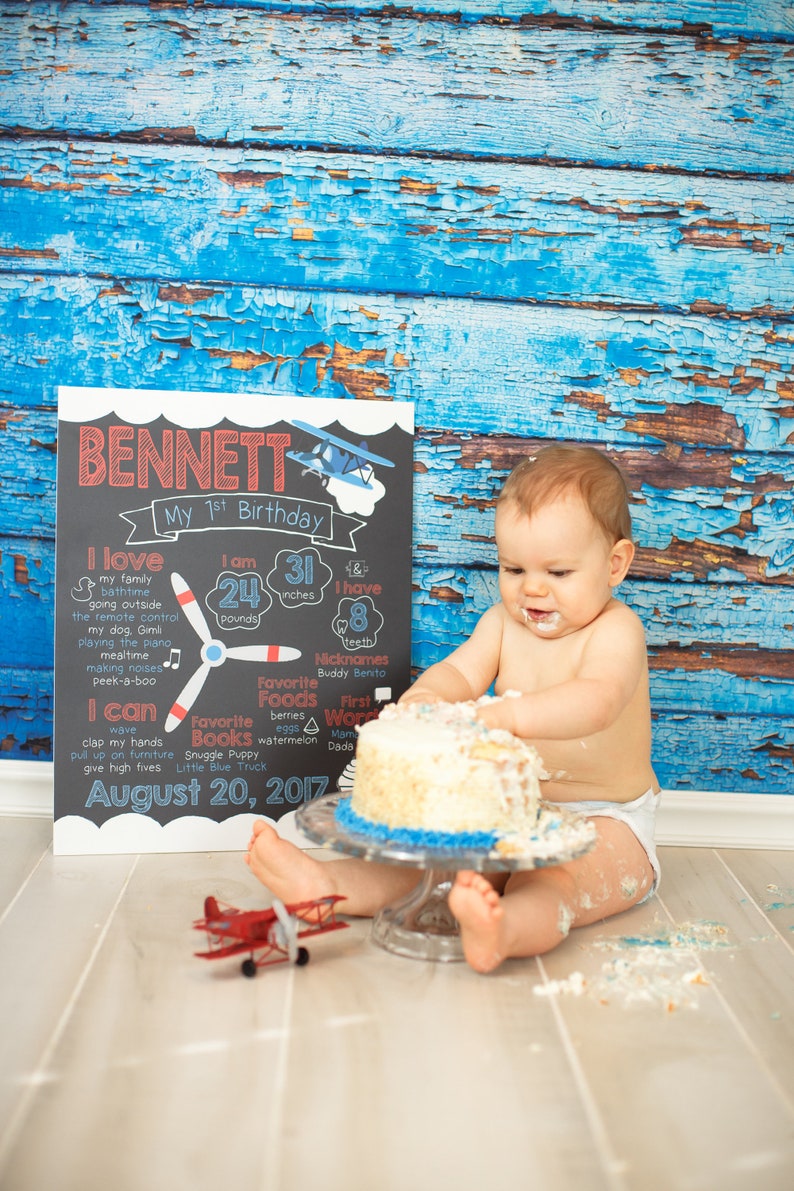 May include: A baby boy sits on a wooden floor with a blue and white cake in front of him. He is wearing a white diaper and has frosting on his face and hands. A chalkboard sign behind him says "Bennett My 1st Birthday" and lists his age, weight, height, and other details. A red toy airplane is on the floor in front of him.
