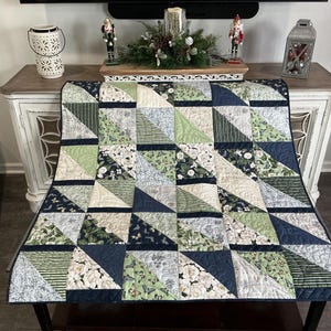 May include: A quilted throw blanket with a geometric pattern. The quilt features triangles in shades of navy, green, and cream, with floral and striped designs. The blanket is displayed on a dark brown table.