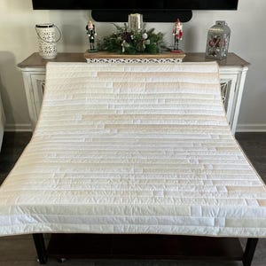 May include: A cream-colored, quilted throw blanket with a rectangular design. The blanket features horizontal stitching and a patchwork pattern of light beige and cream-colored fabric. The blanket is draped over a dark brown bench.