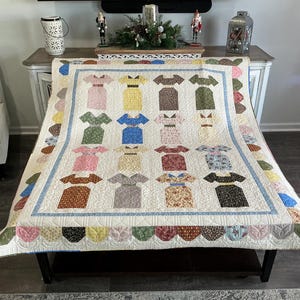 May include: A handmade quilt featuring a repeating pattern of colorful dress shapes. The dresses are in various colors and patterns, including pink, blue, yellow, and brown, set against a white background. The quilt has a scalloped edge and a blue border.