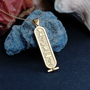 K&ouml;nnte beinhalten: Goldene Halskette mit Anh&auml;nger mit &auml;gyptischem Hieroglyphen-Design. Der Anh&auml;nger ist lang und rechteckig mit einer Reihe von erhabenen Symbolen, darunter ein Vogel, eine Sonne, ein Skarab&auml;us und ein Auge.