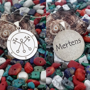 May include: Silver pendant necklace with a circular design featuring a symbol and the letters "M A R S A B". The back of the pendant is engraved with the name "Mertens".