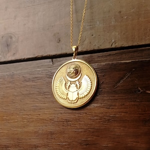 May include: Gold pendant necklace featuring a round charm with an Egyptian scarab beetle design. The charm has a raised scarab beetle with wings and a sun disk with the Eye of Horus above it.