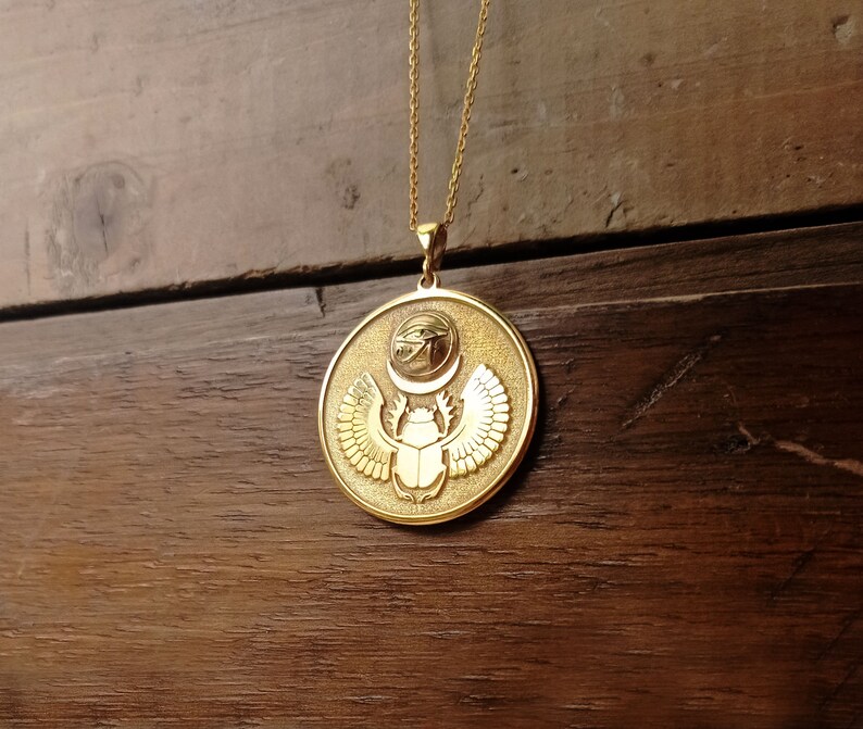 May include: Gold pendant necklace featuring a round charm with an Egyptian scarab beetle design. The charm has a raised scarab beetle with wings and a sun disk with the Eye of Horus above it.