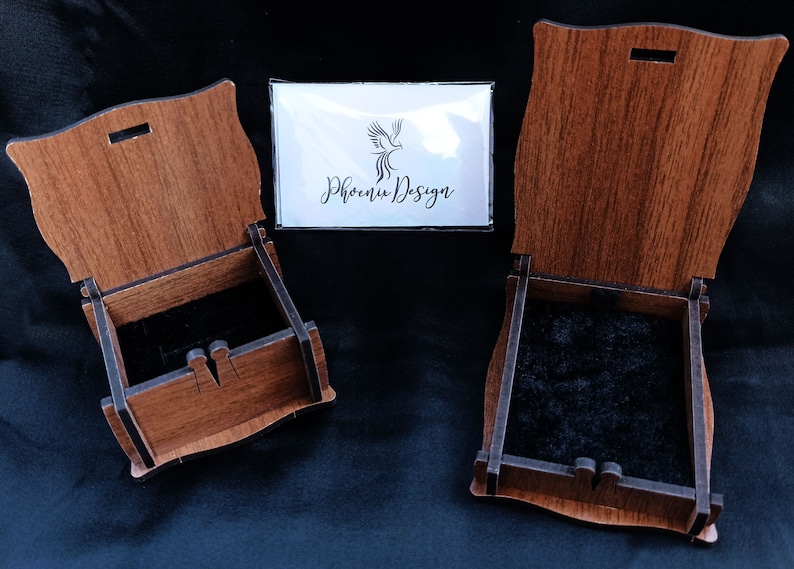 May include: Two wooden boxes with lids open, revealing black velvet interiors. The boxes are made of light brown wood with a dark brown stain. The boxes are both rectangular in shape and have a slightly curved top. The boxes are sitting on a black surface. The text "Phoenix Design" is visible on a white card in the background.