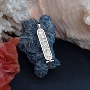 K&ouml;nnte beinhalten: Silberne Halskette mit einem Anh&auml;nger mit &auml;gyptischem Hieroglyphen-Design. Der Anh&auml;nger ist rechteckig mit einer &Ouml;se oben zum Anbringen an einer Kette. Die Hieroglyphen sind in den Anh&auml;nger einge&auml;tzt und umfassen einen Vogel, eine Welle, eine Doppellinie und einen Vogel mit einer Sonnenscheibe.