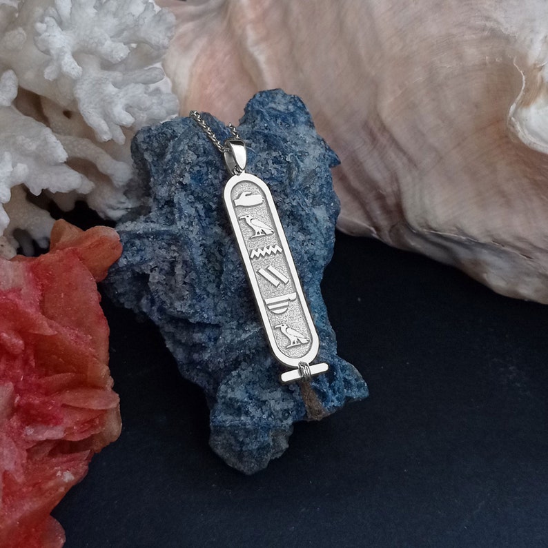 K&ouml;nnte beinhalten: Silberne Halskette mit einem Anh&auml;nger mit &auml;gyptischem Hieroglyphen-Design. Der Anh&auml;nger ist rechteckig mit einer &Ouml;se oben zum Anbringen an einer Kette. Die Hieroglyphen sind in den Anh&auml;nger einge&auml;tzt und umfassen einen Vogel, eine Welle, eine Doppellinie und einen Vogel mit einer Sonnenscheibe.