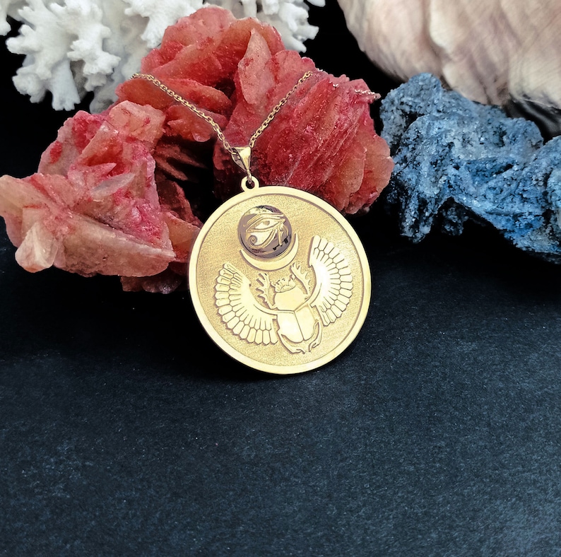 May include: Gold necklace with a round pendant featuring an Egyptian scarab beetle with wings and the Eye of Horus symbol.