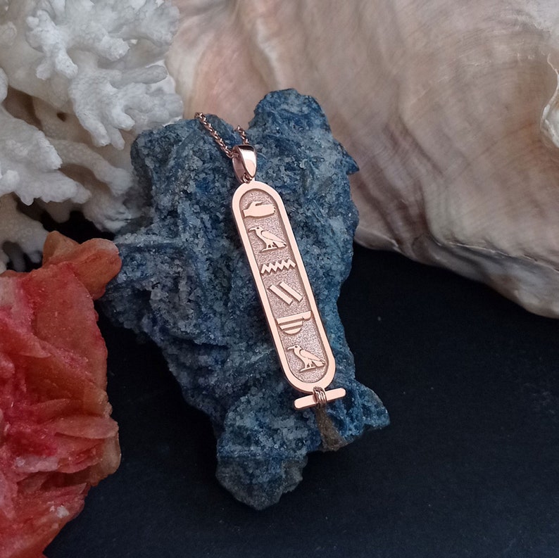 K&ouml;nnte beinhalten: Halskette mit einem Anh&auml;nger aus Ros&eacute;gold, der mit einem &auml;gyptischen Hieroglyphen-Design verziert ist. Der Anh&auml;nger ist rechteckig, hat eine strukturierte Oberfl&auml;che und eine &Ouml;se oben zum Anbringen an einer Kette.