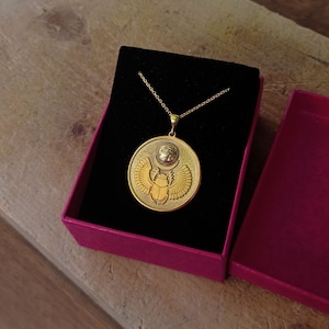 May include: A gold necklace with a round pendant featuring a scarab beetle with outstretched wings and the Eye of Horus above it. The pendant is in a red velvet box.