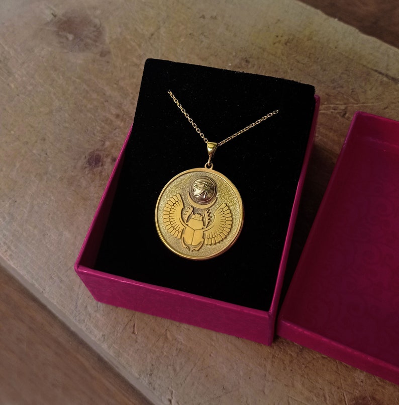 May include: A gold necklace with a round pendant featuring a scarab beetle with outstretched wings and the Eye of Horus above it. The pendant is in a red velvet box.