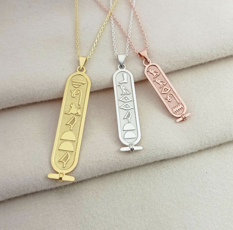 K&ouml;nnte beinhalten: Drei Halsketten aus Gold, Silber und Ros&eacute;gold mit einem gravierten &auml;gyptischen Hieroglyphen-Anh&auml;nger. Jeder Anh&auml;nger hat eine rechteckige Form mit einer Reihe von Hieroglyphen.