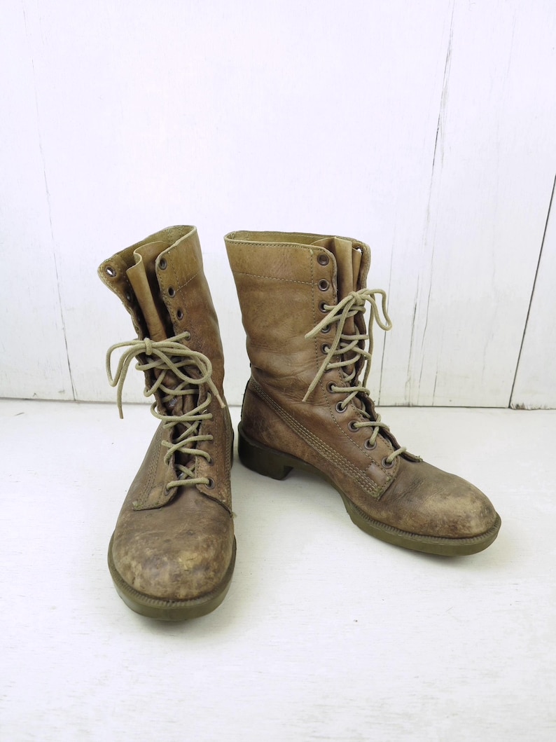 May include: A pair of worn, brown leather combat boots with laces. The boots have a distressed appearance, with visible wear and tear on the leather and soles. The laces are tan, and the boots have a classic, military-inspired design.