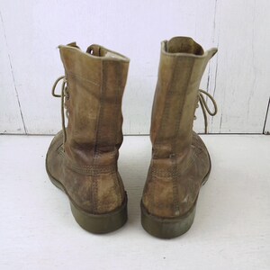 May include: A pair of worn, tan leather combat boots. The boots have laces, a rounded toe, and a green sole. The leather shows signs of wear and tear, with visible creases and discoloration, suggesting they are vintage.