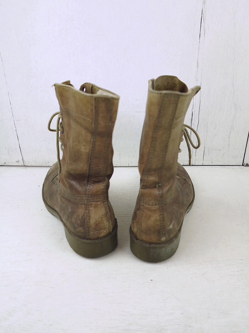 May include: A pair of worn, tan leather combat boots. The boots have laces, a rounded toe, and a green sole. The leather shows signs of wear and tear, with visible creases and discoloration, suggesting they are vintage.