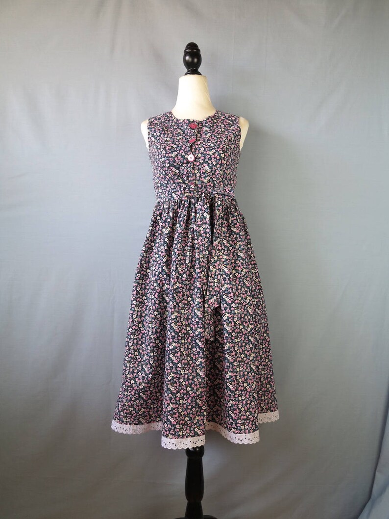 May include: A sleeveless, floral-print dress on a mannequin. The dress features a dark background with a pattern of small pink and white flowers. It has a fitted bodice, a button-up front, and a flared skirt with a lace trim at the hem.