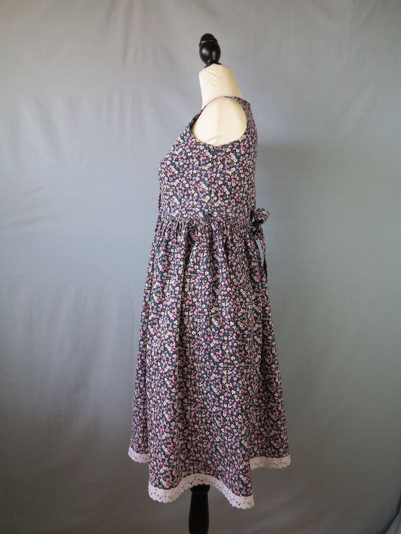 May include: A sleeveless floral print dress with a scoop neckline and a gathered skirt. The dress features a dark navy background with small pink and white flowers. The hemline is trimmed with white lace. A bow detail is visible at the back.