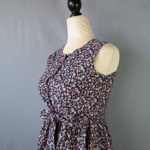 May include: Sleeveless floral print dress with a button-up front and a tie-waist detail. The dress features a navy blue background with a pattern of small pink, white, and blue flowers. The dress is displayed on a mannequin.