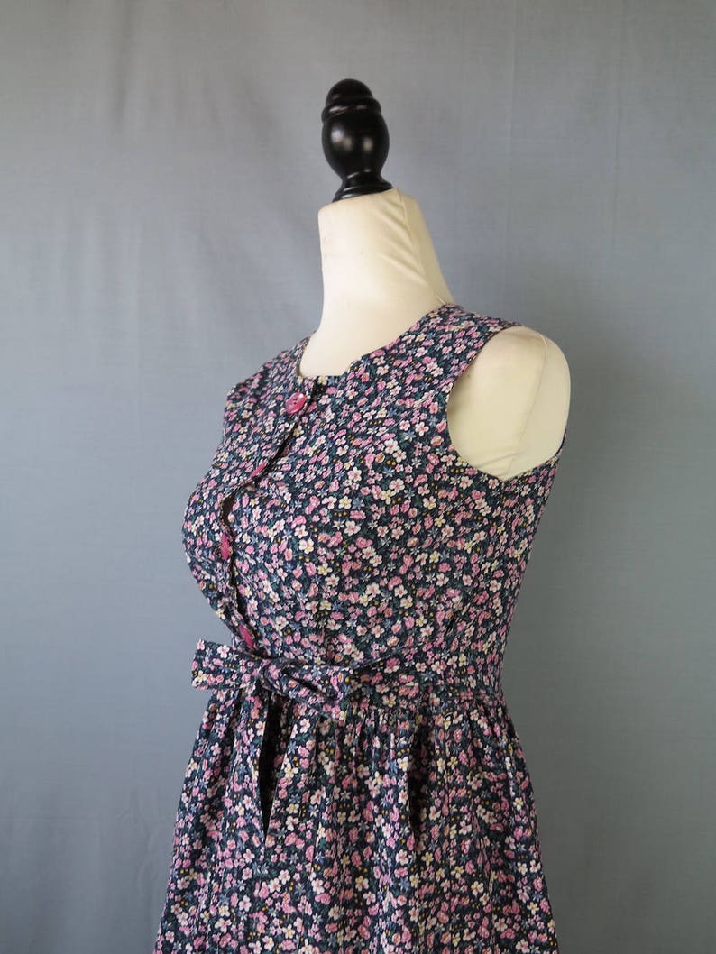 May include: Sleeveless floral print dress with a button-up front and a tie-waist detail. The dress features a navy blue background with a pattern of small pink, white, and blue flowers. The dress is displayed on a mannequin.