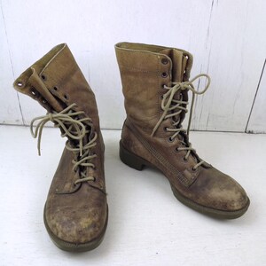 May include: A pair of worn, brown leather combat boots with laces. The boots have a distressed appearance, showing signs of wear and use. The laces are tied, and the boots are set against a white background.