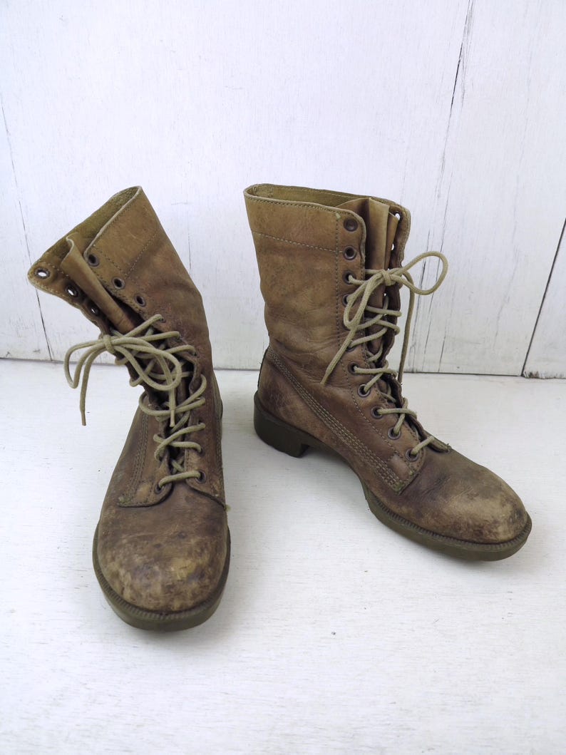 May include: A pair of worn, brown leather combat boots with laces. The boots have a distressed appearance, showing signs of wear and use. The laces are tied, and the boots are set against a white background.