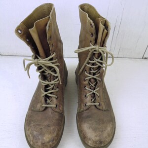May include: A pair of worn, brown leather combat boots. The boots have a lace-up design with multiple eyelets and tan laces. The tops of the boots are folded down, revealing a lighter brown interior. The soles are a darker brown.