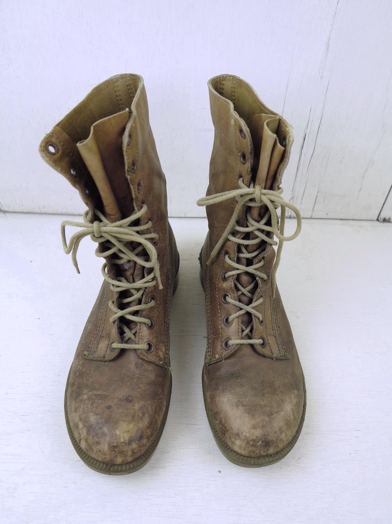 May include: A pair of worn, brown leather combat boots. The boots have a lace-up design with multiple eyelets and tan laces. The tops of the boots are folded down, revealing a lighter brown interior. The soles are a darker brown.