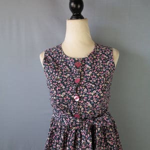 May include: A sleeveless floral print dress with a round neckline and pink buttons. The dress features a fitted bodice and a flared skirt, cinched at the waist with a matching fabric belt. The fabric is a dark navy blue with a pattern of small pink and white flowers.