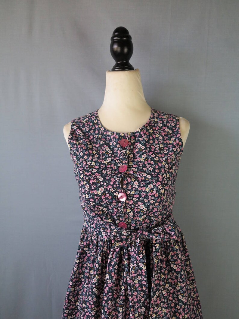 May include: A sleeveless floral print dress with a round neckline and pink buttons. The dress features a fitted bodice and a flared skirt, cinched at the waist with a matching fabric belt. The fabric is a dark navy blue with a pattern of small pink and white flowers.