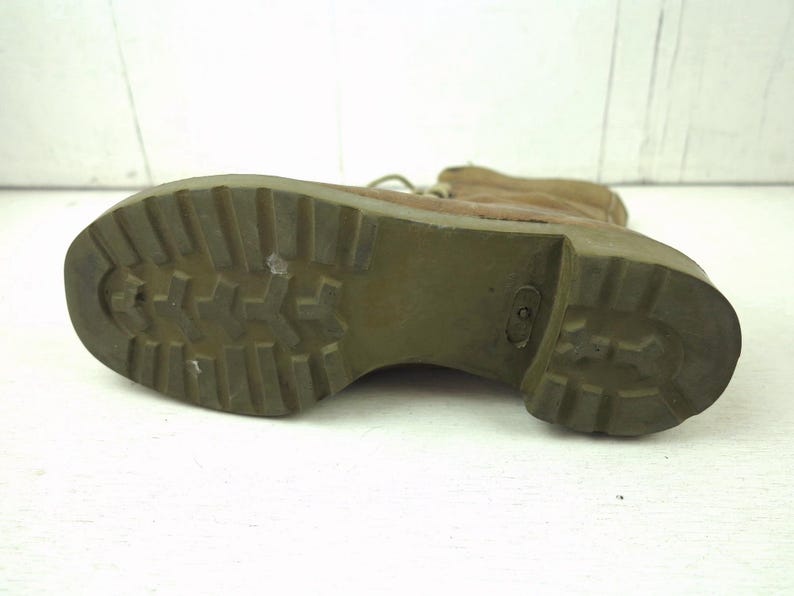 May include: Close-up of the sole of a tan combat boot. The olive green sole has a deep tread pattern for grip. The boot's upper is a light brown color, and the laces are visible. The boot is set against a white background.