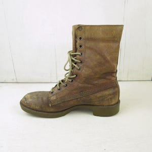May include: A worn, brown leather combat boot with a lace-up design. The boot has a distressed appearance, with visible wear and tear on the leather. The sole is a muted green color, and the laces are a lighter shade of brown.