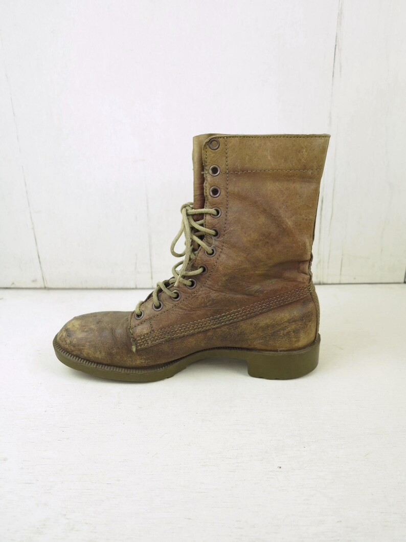 May include: A worn, brown leather combat boot with a lace-up design. The boot has a distressed appearance, with visible wear and tear on the leather. The sole is a muted green color, and the laces are a lighter shade of brown.