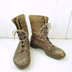 May include: A pair of worn, brown leather combat boots with beige laces. The boots have a distressed appearance, showing signs of wear and use. The soles are a muted green color. The boots are laced up and ready to wear.