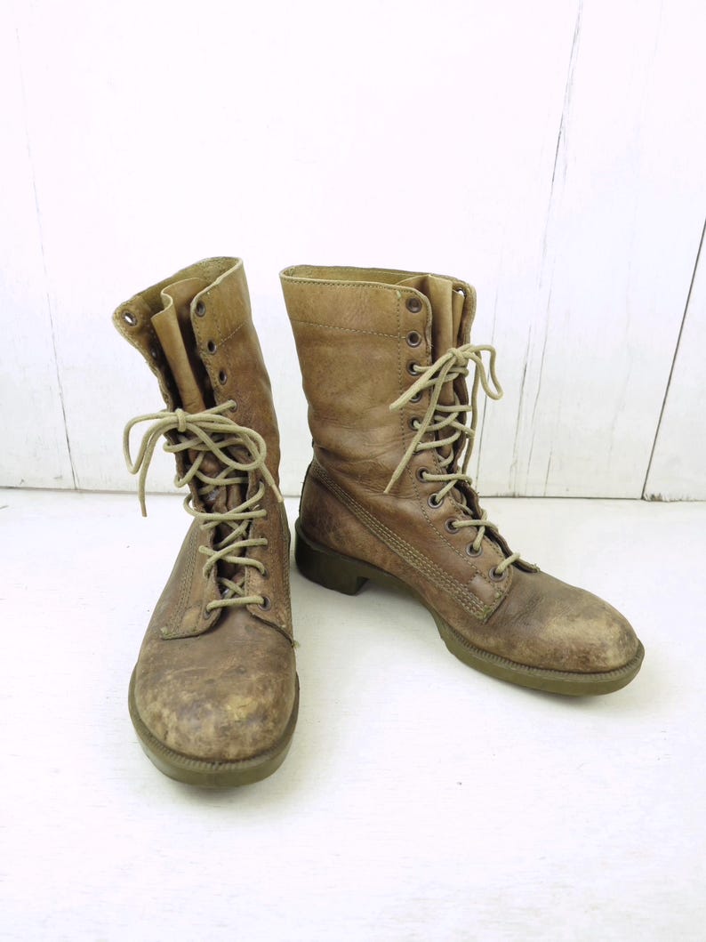 May include: A pair of worn, brown leather combat boots with beige laces. The boots have a distressed appearance, showing signs of wear and use. The soles are a muted green color. The boots are laced up and ready to wear.