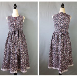 May include: A sleeveless, floral-print dress with a round neckline and a fitted waist. The dress features a dark background with small pink and white flowers. It has a row of buttons down the front and a white lace trim at the hem. The dress is displayed on a mannequin.