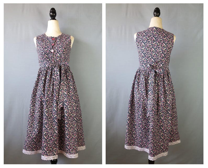 May include: A sleeveless, floral-print dress with a round neckline and a fitted waist. The dress features a dark background with small pink and white flowers. It has a row of buttons down the front and a white lace trim at the hem. The dress is displayed on a mannequin.
