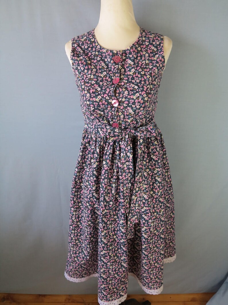 May include: A sleeveless floral dress with a round neckline and pink buttons down the front. The dress features a fitted bodice, a cinched waist, and a flared skirt with a lace trim. The fabric is a dark background with small pink, white, and blue flowers.