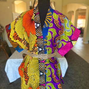 Könnte beinhalten: Eine lebendige, gemusterte Kimono-Jacke mit einem goldenen Gürtel. Die Jacke weist eine Mischung aus kräftigen Farben und Designs auf, darunter Gelb, Orange, Lila und Blau. Der Gürtel sorgt für einen Hauch von Glanz. Das Kleidungsstück wird auf einer schwarzen Schaufensterpuppe präsentiert.