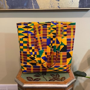 May include: A rectangular tote bag with a vibrant Kente cloth pattern. The bag features a yellow background with a repeating geometric design in green, blue, red, and black. The bag has matching fabric handles.