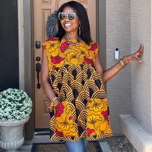 May include: A sleeveless tunic top featuring a vibrant African-inspired print with bold yellow, red, and black floral and geometric patterns. The garment has a round neckline, a gathered waistline, and two large patch pockets on the front.