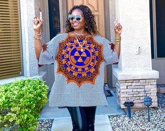 May include: A patterned tunic top with a large circular design in blue and orange. The top has short sleeves and is paired with black leather pants. The outfit is completed with sunglasses and jewelry.