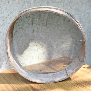 May include: A round wooden flour sifter with a mesh screen. The sifter is made of natural wood and has a rustic appearance.