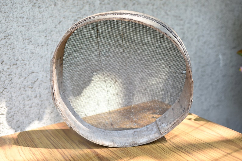 May include: A round wooden flour sifter with a mesh screen. The sifter is made of natural wood and has a rustic appearance.