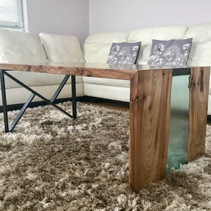 May include: A wooden coffee table with a clear epoxy resin inlay. The table has a black metal X-shaped base and sits on a brown shag rug.