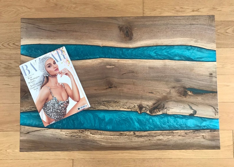 Puede incluir: Una mesa de madera con dos incrustaciones de r&iacute;o de resina azul. Una revista titulada "BAZAAR" est&aacute; encima de la mesa.