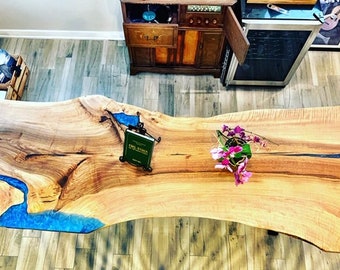 May include: A large, live edge wooden table with a blue epoxy river running through the center. The table has a natural wood finish and is decorated with a small book and a potted plant with pink flowers.