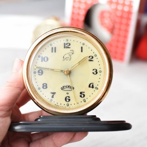 May include: A gold-colored alarm clock with a white face and black numbers. The clock has a small elephant illustration on the face and the text "ОРУМАС" in Cyrillic script. The clock is sitting on a black stand.