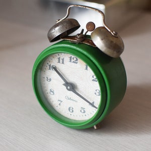 May include: A green alarm clock with silver bells and a white face. The clock has black hands and black numbers. The clock face says "Gebrüder" in the centre.