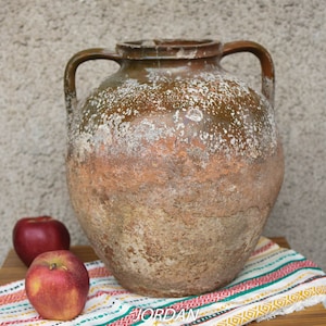 May include: A large, brown, earthenware jug with two handles. The jug has a rough, textured surface and is sitting on a colorful striped table runner.