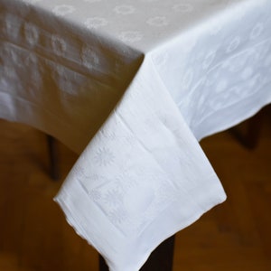 May include: White damask tablecloth with a floral pattern. The tablecloth is folded over a table and the corner is hanging down.